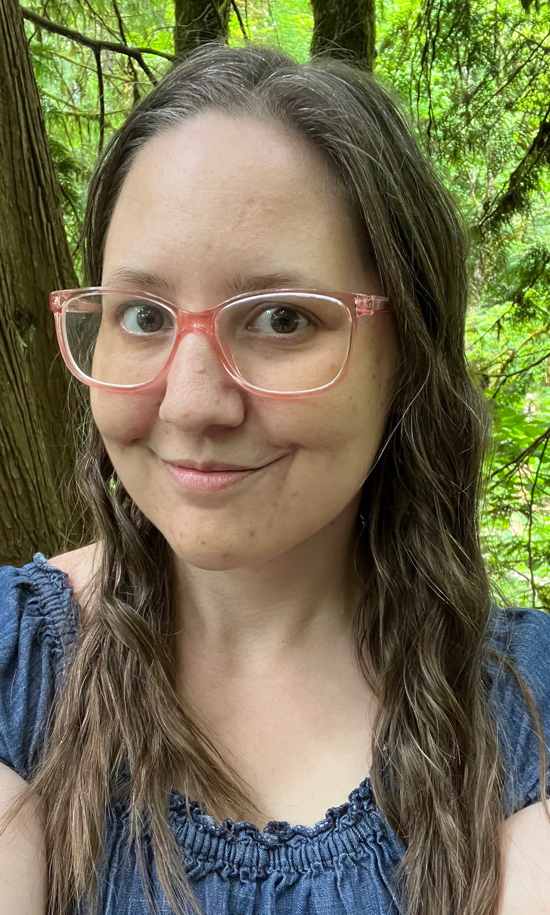 Picture of a woman with long brunette hair wearing a blue dress and pink glasses in a forest.